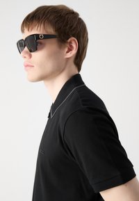 Polo shirt nera con logo discreto, con design a colletto e maniche corte. Occhiali da sole quadrati con montatura nera e lenti scure.