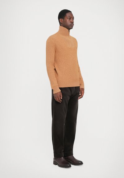 Rjava rebrasta pulover s visokim ovratnikom, v kombinaciji s temnimi hlačami. Gladka tekstura, oprijet kroj in minimalno dekoracijo. Čevlji z gumijastim podplatom.