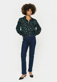 Blusa negra con patrón verde-azul, con botones en el frente y mangas largas, combinada con jeans rectos de color azul oscuro y tacones negros de punta.