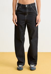 Pantaloni in denim neri con vita alta, gamba larga, cuciture bianche, tasche frontali e chiusura con un singolo bottone. Abbinati a sneakers blu.