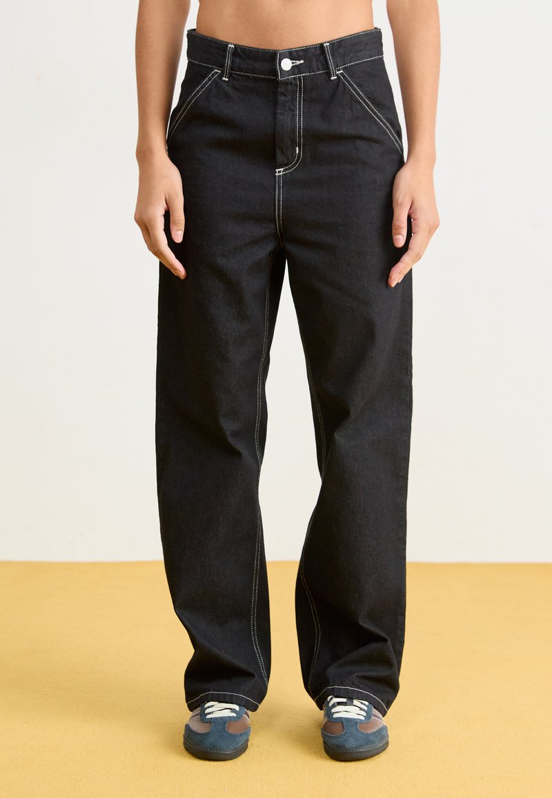 Pantaloni in denim neri con vita alta, gamba larga, cuciture bianche, tasche frontali e chiusura con un singolo bottone. Abbinati a sneakers blu.