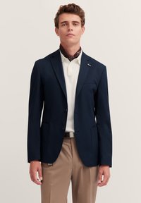 Blazer navy con due bottoni, rever a scudo e tasche frontali, indossato sopra una polo bianca e una sciarpa marrone, abbinato a pantaloni beige.