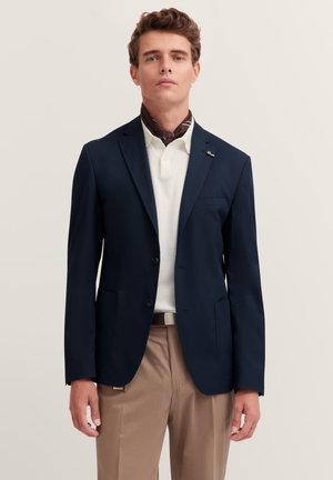 Blazer navy con due bottoni, rever a scudo e tasche frontali, indossato sopra una polo bianca e una sciarpa marrone, abbinato a pantaloni beige.