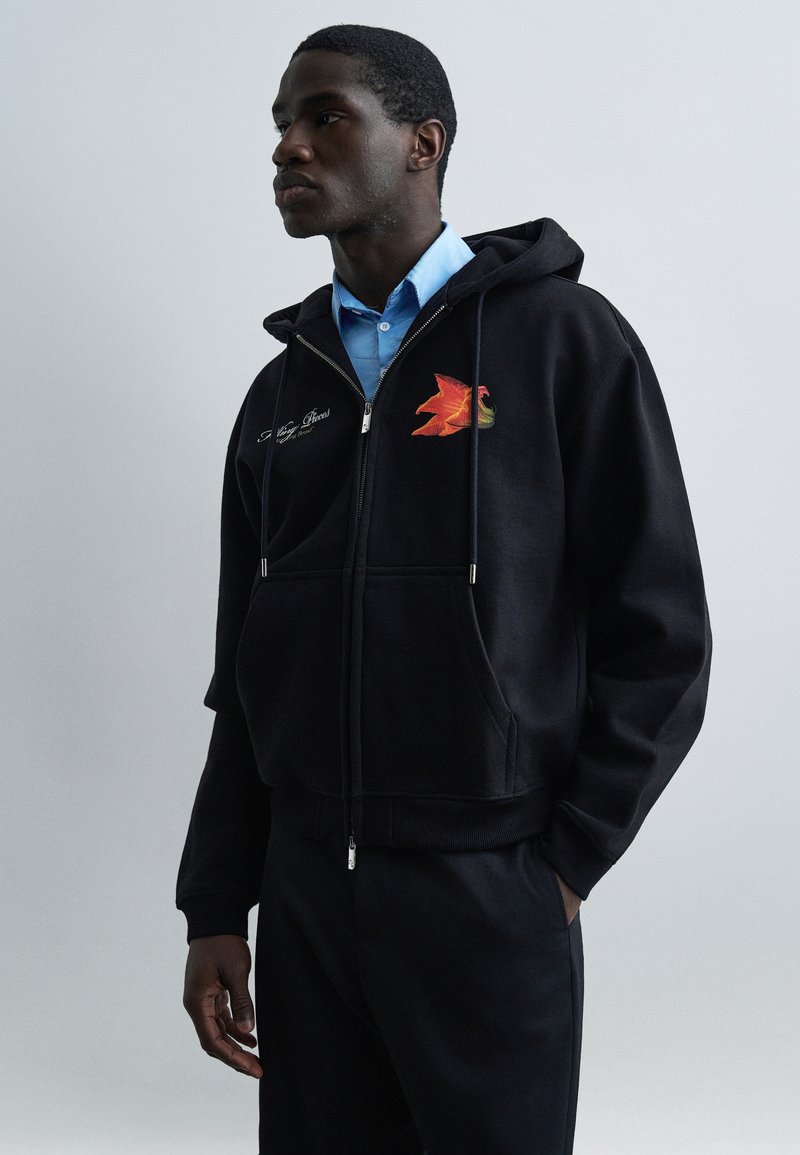 Άνδρας που φοράει μαύρο φούτερ με φερμουάρ και πορτοκαλί σχεδιασμό λουλουδιού, με μπλε πουκάμισο με γιακά από μέσα, στέκεται με το ένα χέρι στην τσέπη μπροστά σε απλό φόντο.