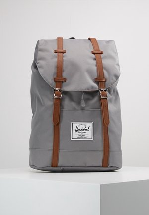 Sac à dos en tissu gris avec des lanières en cuir marron et des boucles argentées, avec un écusson avant portant le nom de la marque sur fond blanc.