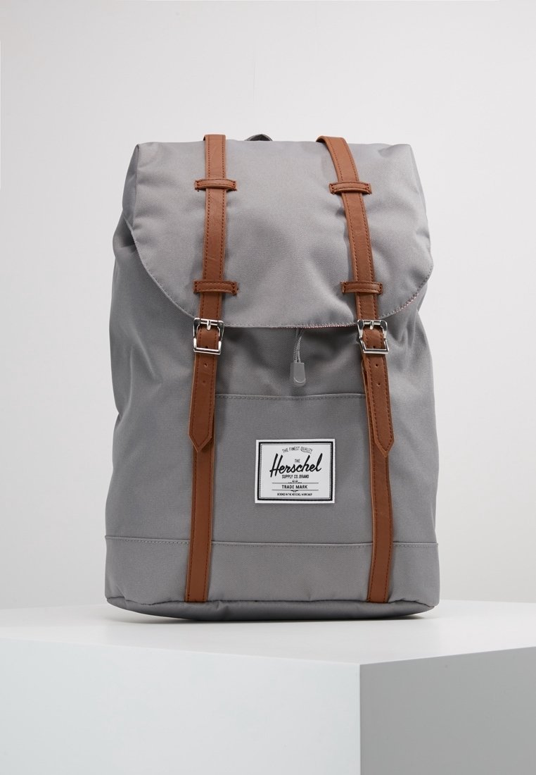 Sac à dos en tissu gris avec des lanières en cuir marron et des boucles argentées, avec un écusson avant portant le nom de la marque sur fond blanc.