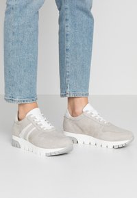 Harmaat mokkanahkaiset tennarit valkoisilla yksityiskohdilla, kuten nauhoilla ja kolmella raidalla. Kenkäpäällä on tekstuuri ja paksu valkoinen kumipohja.