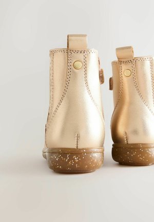 Stivali alla caviglia metallici oro con suole piatte, cuciture laterali, linguette per l'espulsione e suola marrone macchiata. Texture liscia con forma arrotondata.