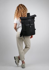 Zaino a rotolo nero realizzato in materiale resistente, dotato di spallacci regolabili, una tasca anteriore con zip e un logo distintivo sulla parte anteriore.