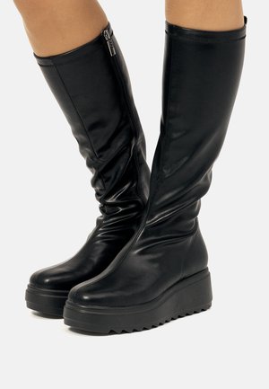 Botas negras hasta la rodilla hechas de cuero liso con un diseño arrugado, que cuentan con un cierre de cremallera lateral y una suela de plataforma robusta.