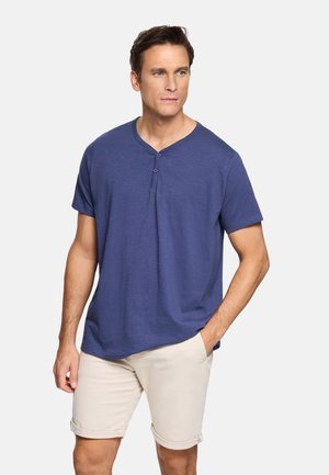 Hombre con camiseta henley de manga corta azul y pantalones cortos beige, de pie con una mano en el bolsillo contra un fondo liso.