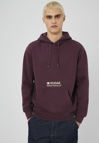 Jeune personne avec des cheveux blonds courts portant un sweat à capuche Kodak violet avec une poche avant et un jean bleu foncé, se tenant contre un fond uni.