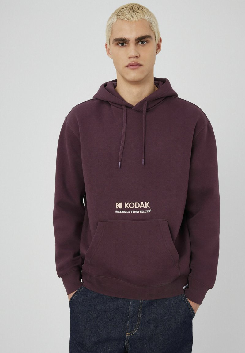 Jeune personne avec des cheveux blonds courts portant un sweat à capuche Kodak violet avec une poche avant et un jean bleu foncé, se tenant contre un fond uni.