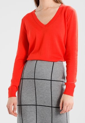 Femme portant un pull rouge vif à col en V associé à une jupe grise taille haute avec un motif à carreaux noirs.