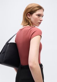 Γυναίκα με κοντά κόκκινα μαλλιά που φοράει ένα ριχτό πουκάμισο σε χρώμα σκουριάς και μαύρο παντελόνι, κρατώντας μια μεγάλη μαύρη δερμάτινη τσάντα ώμου.