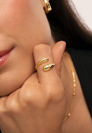 Anillo de oro con un diseño fluidamente alargado y acentos suaves y redondeados. Se lleva puesto en un dedo, destacando el brillo y la textura.