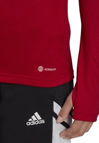 Rode sportieve longsleeve shirt met geribbelde textuur en AEROREADY-logo dicht bij de zoom, gecombineerd met zwarte shorts met witte strepen en het Adidas-logo.