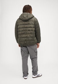 Olivgrüne Pufferjacke mit horizontalen Steppnähten, abnehmbarer Kapuze, kombiniert mit grauen Cargo-Hosen und weißen Sneakern.
