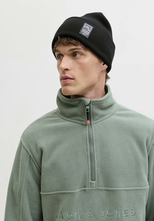 Jonge man die een zwarte beanie en een lichtgroene fleecejas met halve ritssluiting draagt, kijkt naar links tegen een effen achtergrond.