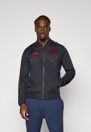 Uomo in piedi che indossa una giacca nera e rossa con zip, con loghi di club, e pantaloni blu navy, davanti a uno sfondo chiaro e uniforme.