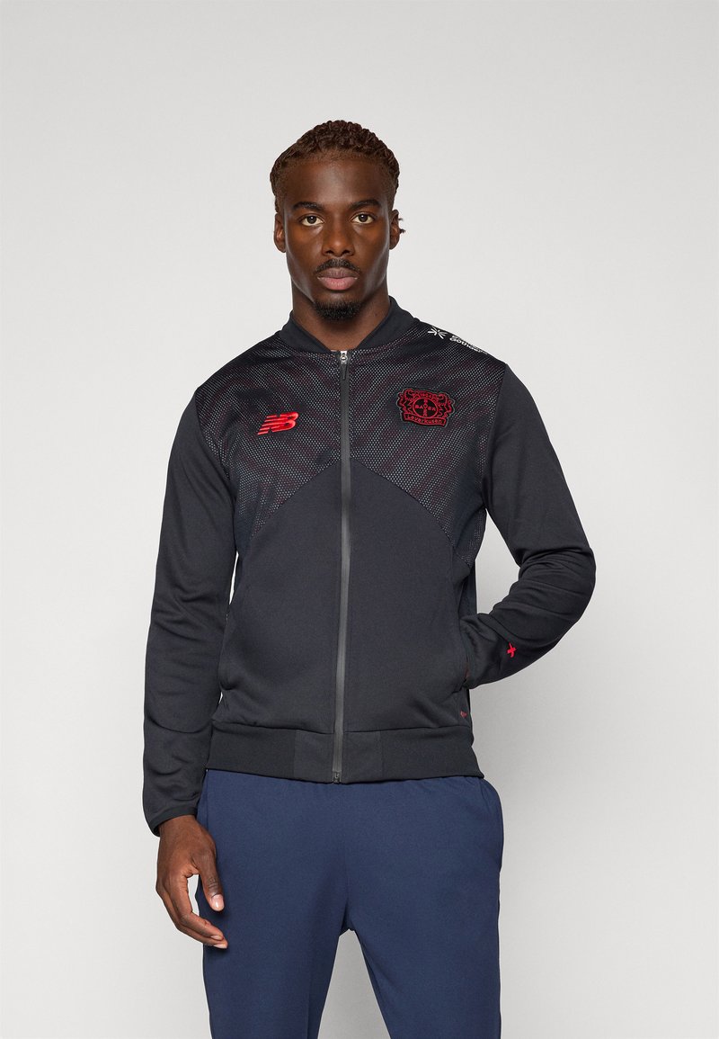 Man står klädd i en svart och röd zip-up jacka med klubbens logotyper och marinblå byxor mot en enkel ljus bakgrund.