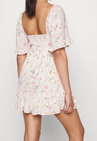 Vestido floral con mangas cortas abullonadas, espalda elástica y dobladillo con volantes. Presenta patrones de flores en tonos rosa pálido y verde sobre un fondo claro.