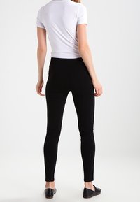 Svarta leggings med en slät yta och en åtsittande design, kombinerat med en vit kortärmad topp och svarta platta skor. Bakifrån.
