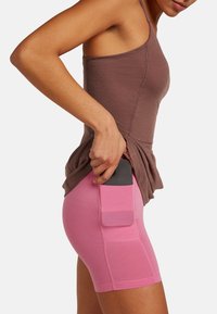 Aansluitende korte broek in lichtroze met een zijzak. Bruine tanktop. Telefoon gedeeltelijk in de zak gestoken. Zachte, rekbare stof.
