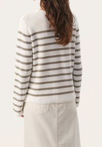 Pitkähiuksinen nainen, jolla on päällään valkoinen neule, jossa harmaat vaakaraitoja, ja kermanvärinen hame, kasvojaan kameraan päin kääntyneenä.