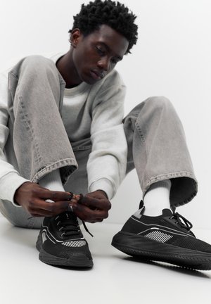 Jeune homme en jean gris et chaussettes blanches assis et attachant des baskets noires sur un fond blanc uni.
