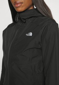 Chaqueta impermeable negra con cierre de cremallera y cuello alto; cuenta con un logotipo blanco contrastante en el lado izquierdo del pecho y una textura suave.