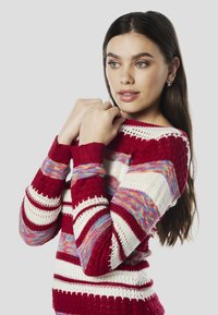 Gebreide trui met rode, witte en meerkleurige horizontale strepen en een gestructureerd patroon. Geribbelde manchetten en opengewerkt detail bij de halslijn.