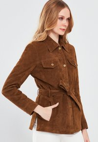 Bruine suède jas met een kraag, knoopsluiting aan de voorkant, twee borstzakken en een gestrikt riem in de taille. Heeft lange mouwen en een gestructureerde afwerking.