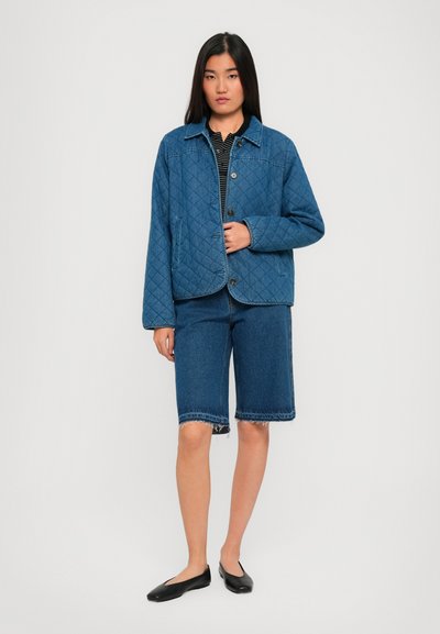 Femeie în picioare, îmbrăcată cu o jachetă matlasată albastră, un tricou cu dungi negre, pantaloni scurți din denim până la genunchi cu margini desfăcute și pantofi negri fără toc.