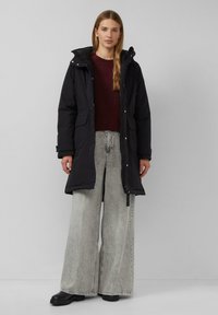 Schwarze Parkajacke mit Kapuze, ausgestattet mit zwei Fronttaschen, kombiniert mit übergroßen grauen Denim-Hosen und schwarzen Stiefeletten.