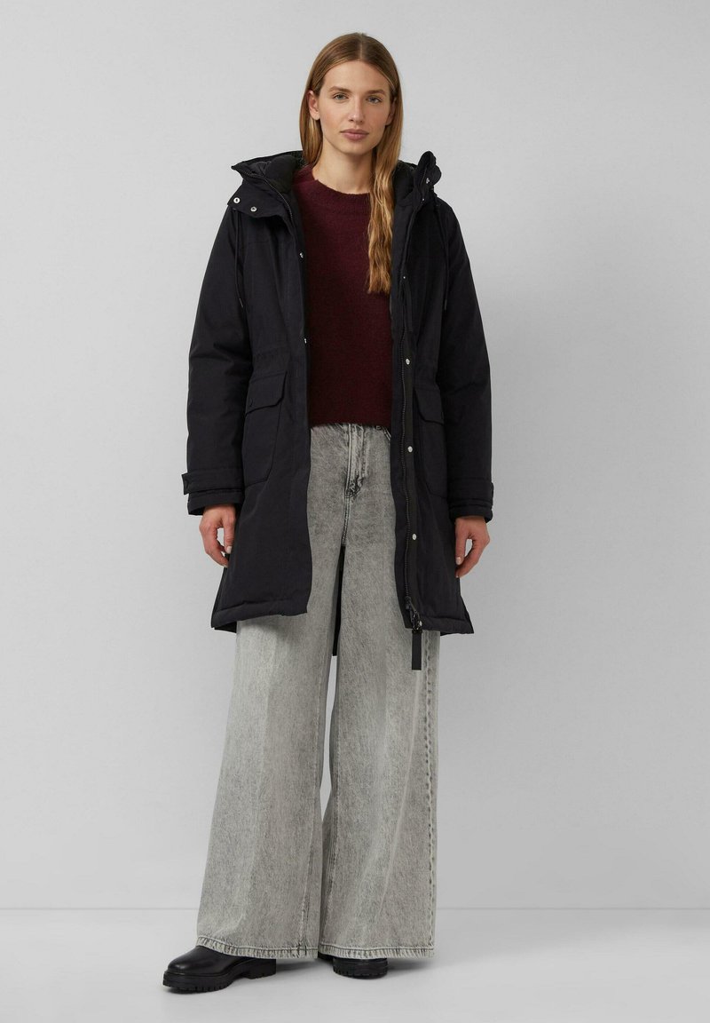 Schwarze Parkajacke mit Kapuze, ausgestattet mit zwei Fronttaschen, kombiniert mit übergroßen grauen Denim-Hosen und schwarzen Stiefeletten.
