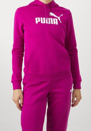 Žena v jasně růžové mikině Puma s přední kapsou a sladěnými tepláky na jednobarevném pozadí.