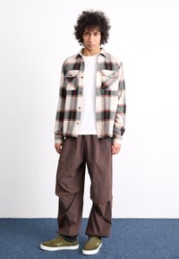 Pārbaudīta flaneļa krekls bēšā, melnā un oranžā krāsā ar divām krūšu kabatām; apvienots ar brīviem brūniem biksēm un zaļām kedām.