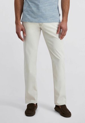 Mann trägt hellbeige, gerade geschnittene Hosen, braune Loafers und ein hellblau meliertes Kurzarmhemd, steht vor einem neutralen Hintergrund.