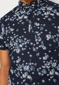 Camicia a maniche corte con bottoni in tessuto blu scuro, caratterizzata da un motivo floreale bianco e blu. Include una tasca sul petto e bottoni neri.