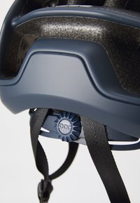 Kask o matowej, ciemnoniebieskiej powłoce, z teksturowanymi akcentami z włókna węglowego, regulowanym pokrętłem z tyłu oraz czarnymi paskami zapewniającymi pewne dopasowanie.