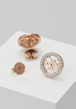 Oorbellen van roségoud met een rond ontwerp, waarvan één met heldere kristallen rond een vierbooglogo op een witte ondergrond.