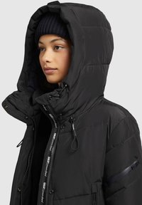Schwarze Pufferjacke mit großer Kapuze, Vorderreißverschluss, seitlichen Taschen und verstellbaren Akzenten. Glatte, gesteppten Textur mit sichtbarem Branding.