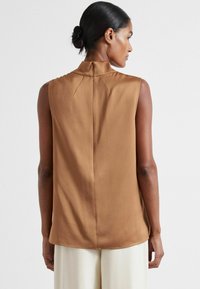 Sleeveless brown top with a high collar, smooth texture, back seam detail, and rounded hem, paired with cream-colored trousers.