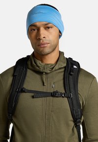 Ein blauer Stoff-Haarband, das über einer dunkelgrünen Zip-Jacke getragen wird, ergänzt durch einen schwarzen Rucksack mit verstellbaren Trägern.