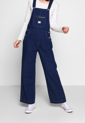 Salopette en denim bleu foncé à jambes larges, équipée de bretelles ajustables, d'une poche avant zippée et de boutons-pression sur les côtés.