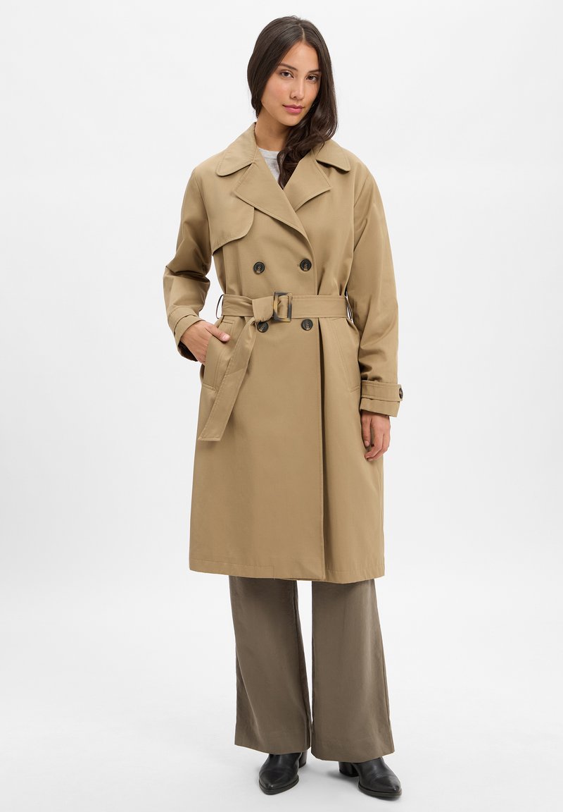 Beiger Trenchcoat mit doppelter Knopfleiste, weitem Kragen und tailliertem Gürtel. Er verfügt über große Knöpfe und seitliche Taschen.