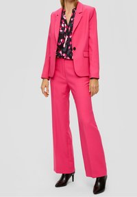 Mujer vestida con blazer rosa brillante y pantalones anchos a juego, con botas negras de tacón alto y una blusa negra con estampados rosas.