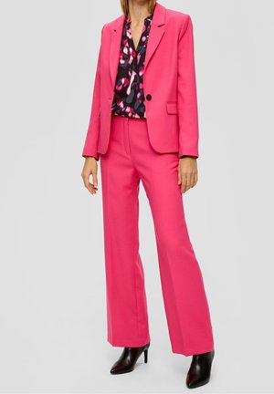 Nainen, joka pukeutuu kirkkaanpunaiseen bleiseriin ja yhteensopiviin leveälahkeisiin housuihin, mustiin korkokenkiin ja mustaan puseroon, jossa on vaaleanpunaisia kuvioita.