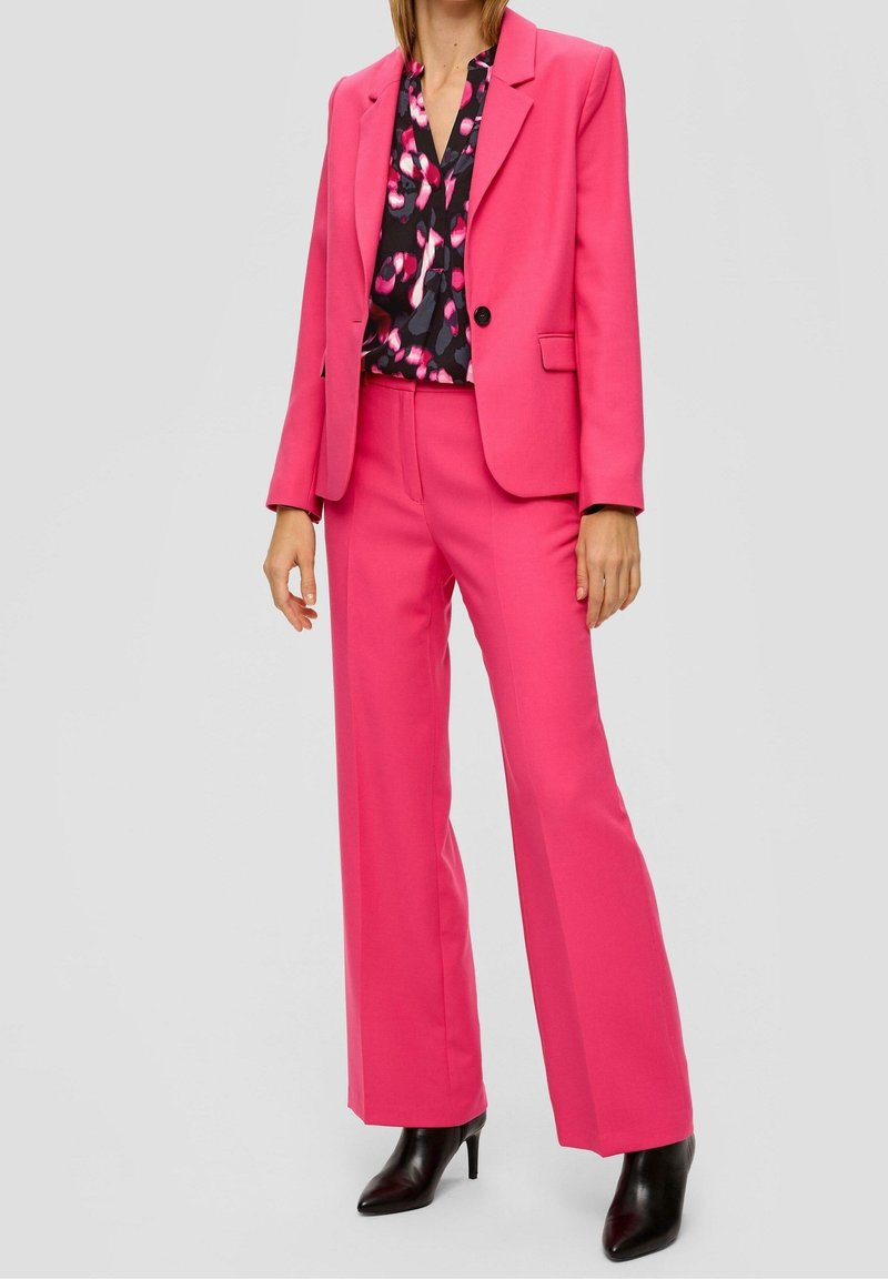 Mujer vestida con blazer rosa brillante y pantalones anchos a juego, con botas negras de tacón alto y una blusa negra con estampados rosas.